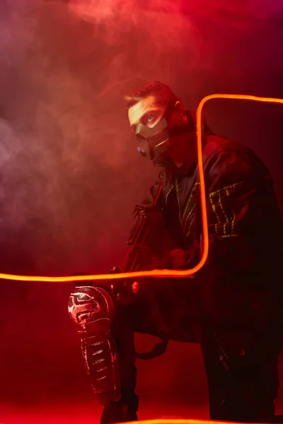 Peligroso jugador bi-racial cyberpunk en máscara protectora sosteniendo pistola cerca de la iluminación de neón en negro con humo - foto de stock