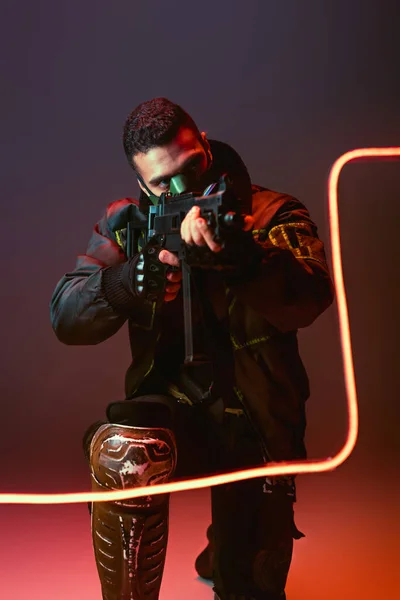Bi-racial cyberpunk hombre en máscara apuntando pistola cerca de neón iluminación en negro - foto de stock