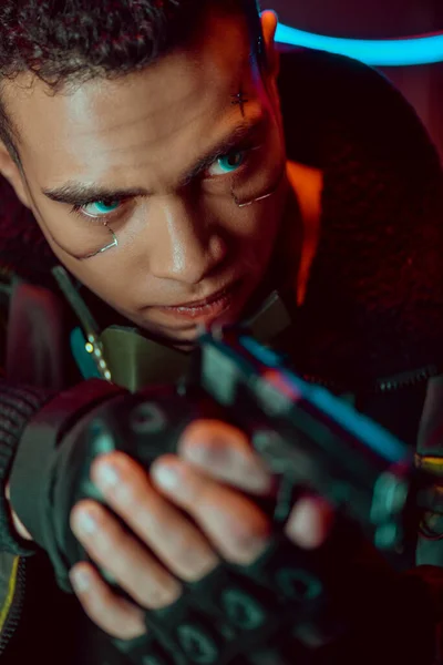 Selective focus of armed bi-racial cyberpunk player with metallic plates on face holding gun — Stock Photo