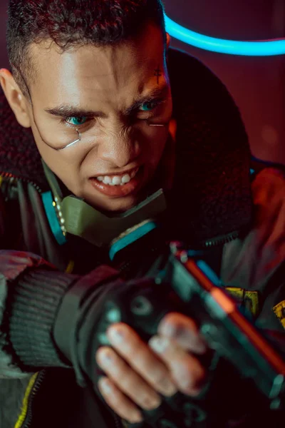Foco seletivo de jogador de cyberpunk bi-racial irritado com placas metálicas no rosto segurando arma — Fotografia de Stock