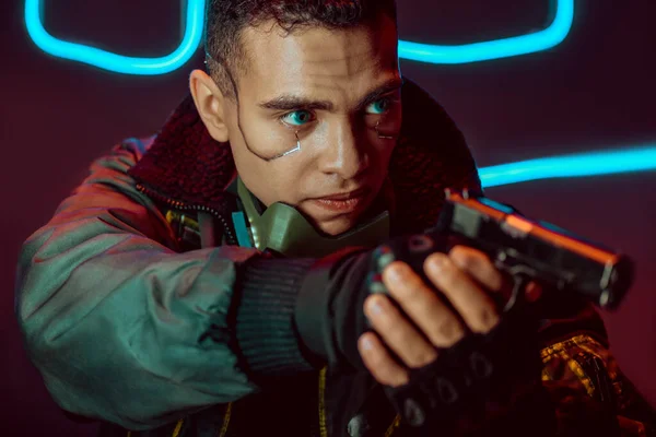 Selective focus of dangerous bi-racial cyberpunk player with gun on black with neon lighting — Stock Photo