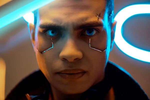 Lecteur cyberpunk bi-racial avec plaques métalliques sur le visage regardant la caméra près de l'éclairage au néon — Photo de stock