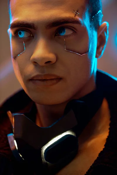 Jugador bi-racial cyberpunk con placas metálicas en la cara mirando hacia otro lado cerca de la iluminación de neón - foto de stock