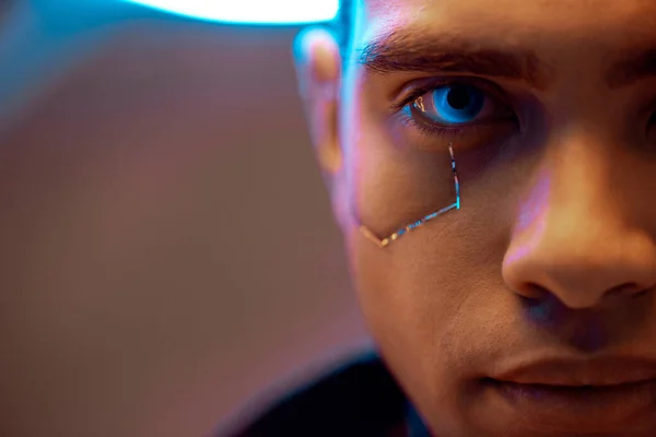 Vista recortada del jugador cyberpunk de raza mixta con placas metálicas en la cara y ojos azules mirando a la cámara - foto de stock