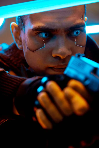 Selective focus of bi-racial cyberpunk player with metallic plates on face and blue eyes holding gun near neon lighting — Stock Photo