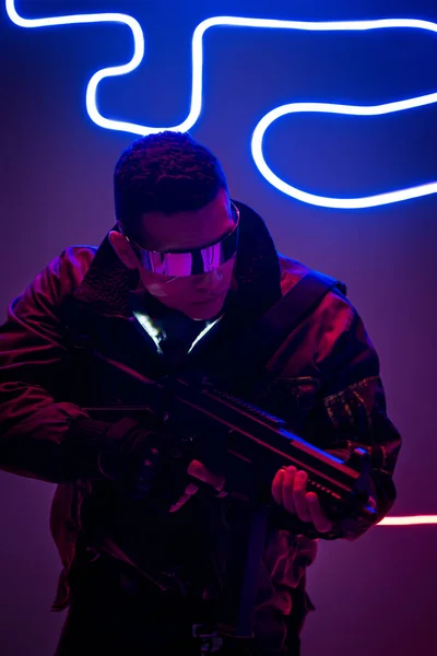 Armed mixed race cyberpunk player in futuristic glasses holding gun near neon lighting — Stock Photo