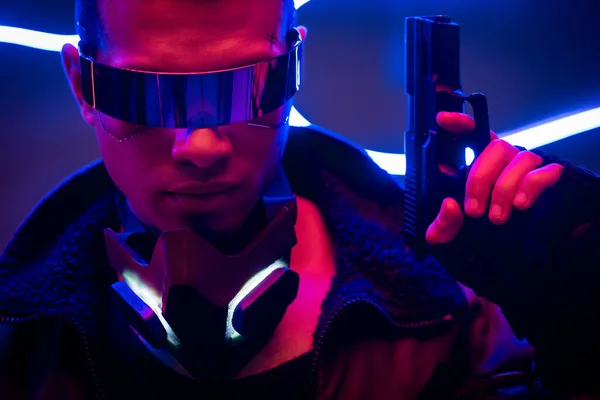 Bel giocatore cyberpunk razza mista in occhiali futuristici con pistola vicino illuminazione al neon blu — Foto stock