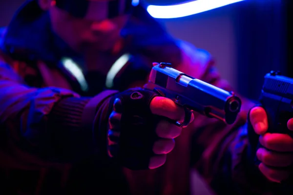 Foco seletivo de jogador de cyberpunk bi-racial segurando armas perto de iluminação de néon — Fotografia de Stock