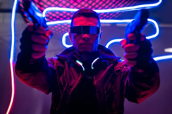Selective focus of armed mixed race cyberpunk player in futuristic glasses holding guns near neon lighting — Stock Photo