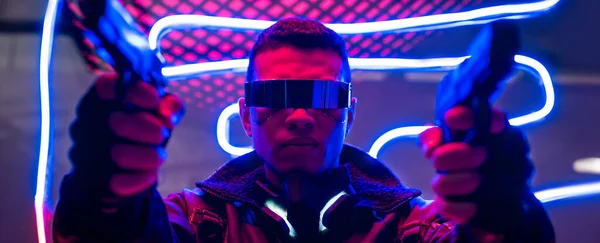 Panoramic shot of mixed race cyberpunk player in futuristic glasses holding guns near neon lighting — Stock Photo