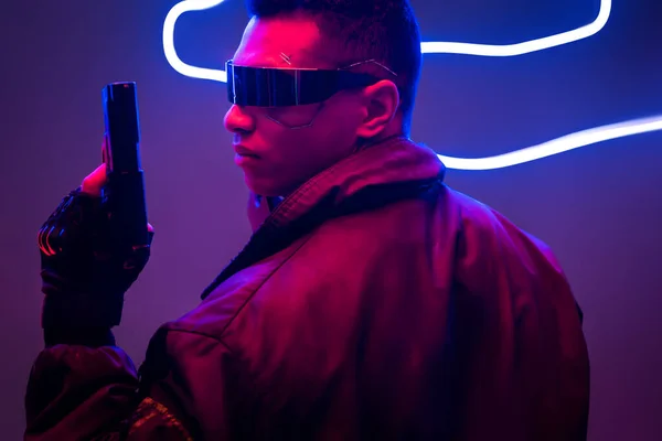 Seitenansicht eines Mixed Race Cyberpunk-Spielers in futuristischer Brille mit Pistole in der Nähe von Neonbeleuchtung — Stockfoto