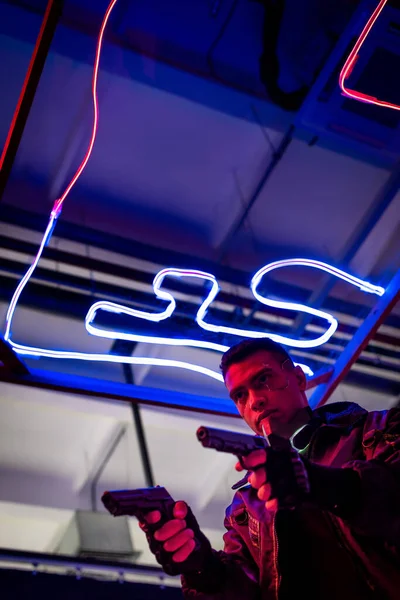 Vista basso angolo di armato giocatore cyberpunk bi-razziale in possesso di pistole vicino illuminazione al neon — Foto stock