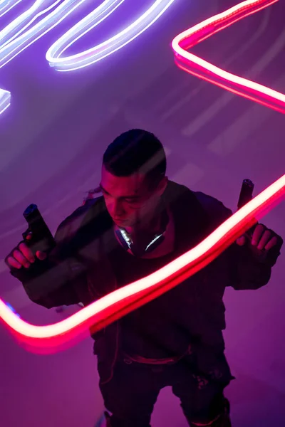 Vista aérea del jugador de cyberpunk bi-racial armado sosteniendo armas cerca de la iluminación de neón - foto de stock