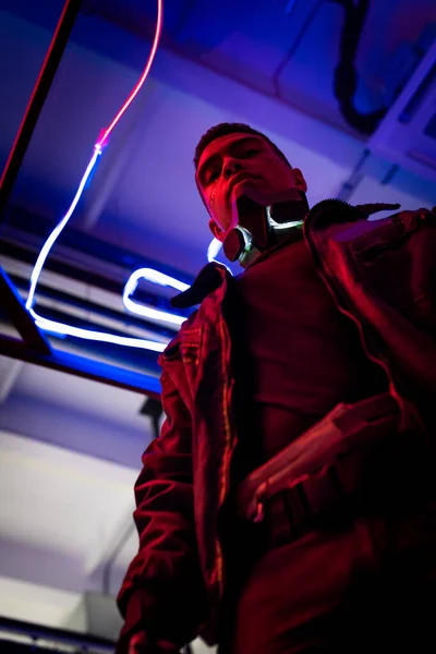 Vista de ángulo bajo del jugador bi-racial cyberpunk de pie cerca de la iluminación de neón azul - foto de stock