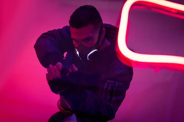 Overhead view of armed bi-racial cyberpunk player in mask holding gun near neon lighting — Stock Photo