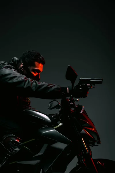 Side view of armed bi-racial cyberpunk player in mask riding motorcycle and holding gun on grey — Stock Photo