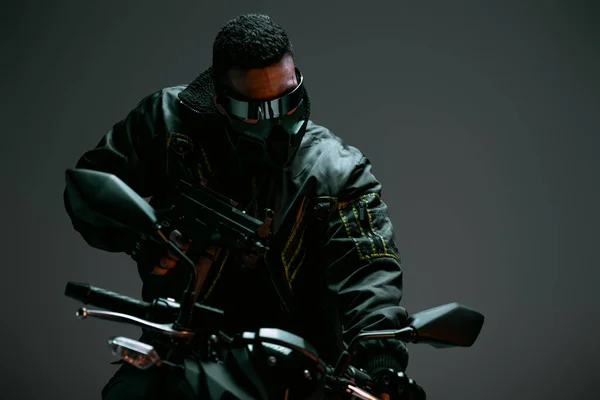 Selective focus of armed bi-racial cyberpunk player in mask and futuristic glasses holding gun and riding motorcycle on grey — Stock Photo