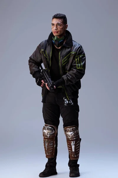 Guapo bi-racial cyberpunk jugador sosteniendo arma y de pie en gris - foto de stock