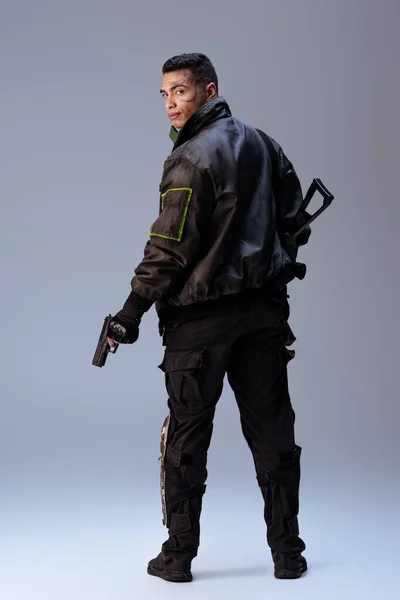 Jugador cyberpunk de raza mixta sosteniendo arma y de pie sobre gris - foto de stock
