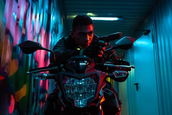 Bi-racial cyberpunk player on motorcycle aiming gun on street with graffiti — Stock Photo