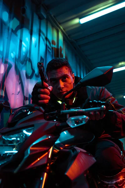 Selective focus of mixed race cyberpunk player on motorcycle aiming gun on street with graffiti — Stock Photo