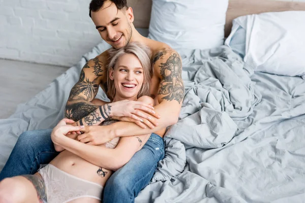 Schöner tätowierter Mann umarmt lächelnde Freundin in Dessous auf dem Bett — Stockfoto