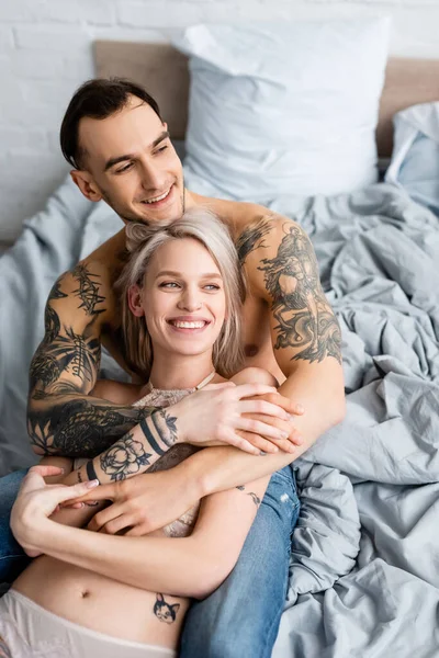 Улыбающийся мужчина обнимает татуированную девушку в нижнем белье на кровати — стоковое фото