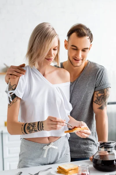 Bel homme tatoué embrassant petite amie avec toast près de la cafetière sur la table dans la cuisine — Photo de stock