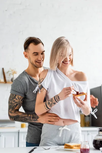 Hombre tatuado abrazando novia sonriente con tostadas y mermelada cerca de la mesa de la cocina - foto de stock