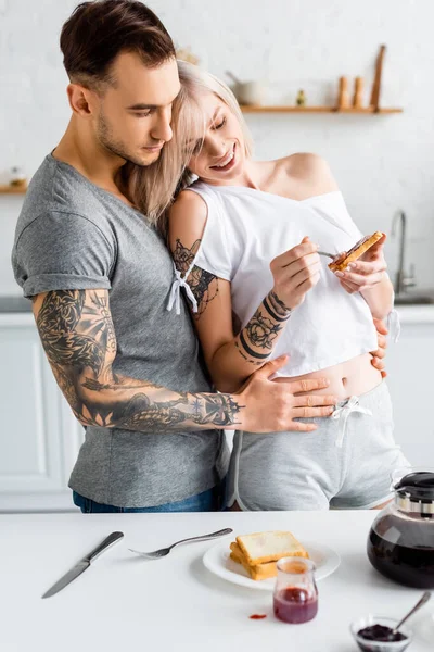 Hombre guapo tatuado abrazando novia sonriente con tostadas cerca de cafetera y mermeladas en la mesa de la cocina - foto de stock