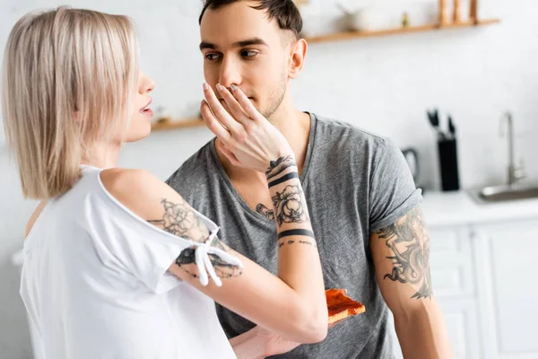 Tattooed woman holding toast and touching face of handsome boyfriend on kitchen — Stock Photo