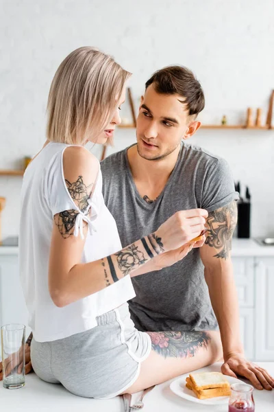 Tattooed man looking at beautiful girlfriend with toast and jam during breakfast in kitchen — Stock Photo