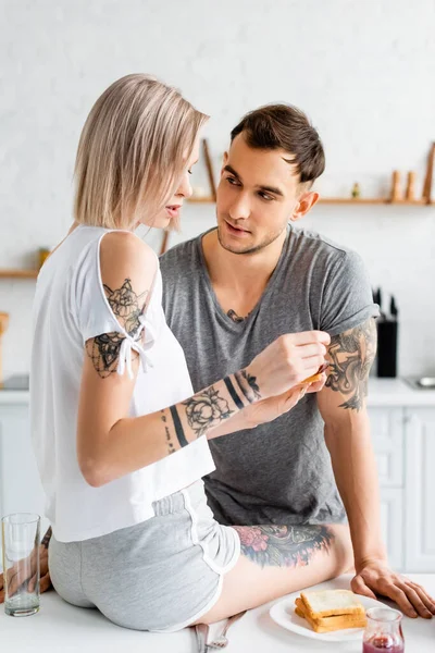 Tatoué homme regardant belle petite amie avec toast et confiture pendant le petit déjeuner dans la cuisine — Photo de stock