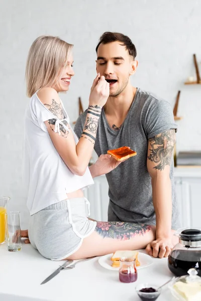 Smiling woman sitting on kitchen table and feeding tattooed boyfriend with jam during breakfast in kitchen — Stock Photo