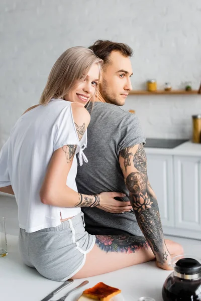 Hermosa chica sonriendo a la cámara mientras abraza novio tatuado durante el desayuno en la cocina - foto de stock