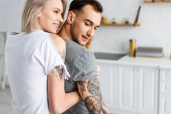 Vista posteriore della donna sorridente che abbraccia il fidanzato tatuato in cucina — Foto stock