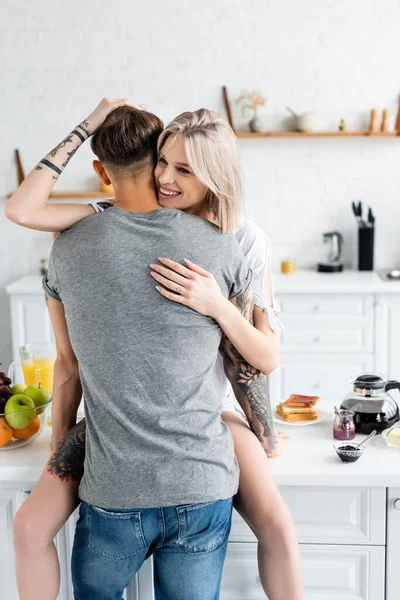 Mujer sonriente abrazando novio tatuado cerca del desayuno en la mesa de la cocina - foto de stock