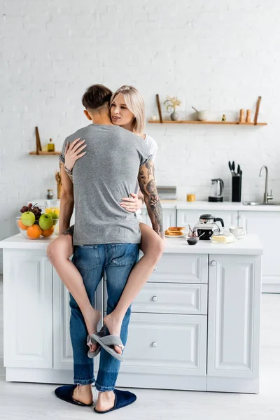 Homme tatoué embrassant belle petite amie près du café et des toasts sur la table de cuisine — Photo de stock