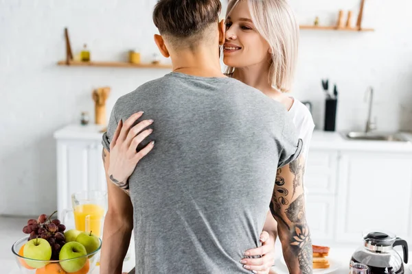 Hermosa chica sonriente abrazando novio tatuado cerca del desayuno en la mesa de la cocina - foto de stock