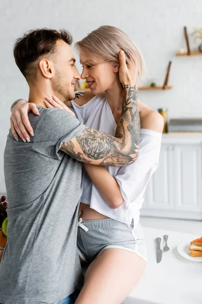 Vista laterale dell'uomo tatuato che abbraccia la fidanzata sorridente sul tavolo della cucina — Foto stock