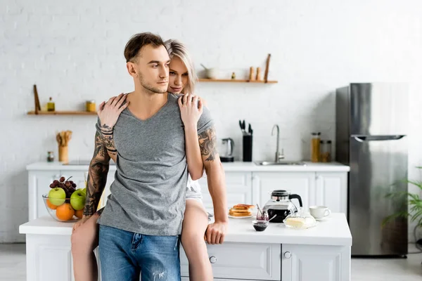 Hermosa mujer abrazando novio tatuado cerca del desayuno en la mesa de la cocina - foto de stock