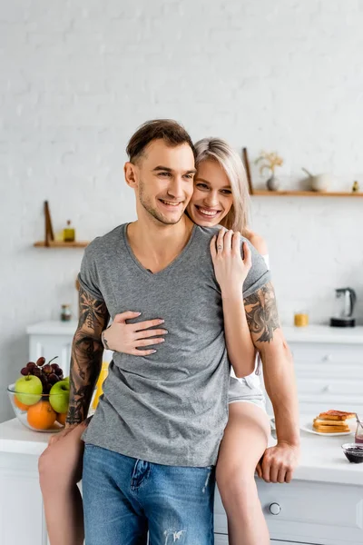 Belle femme étreignant petit ami tatoué souriant près des toasts et des fruits sur la table de cuisine — Photo de stock