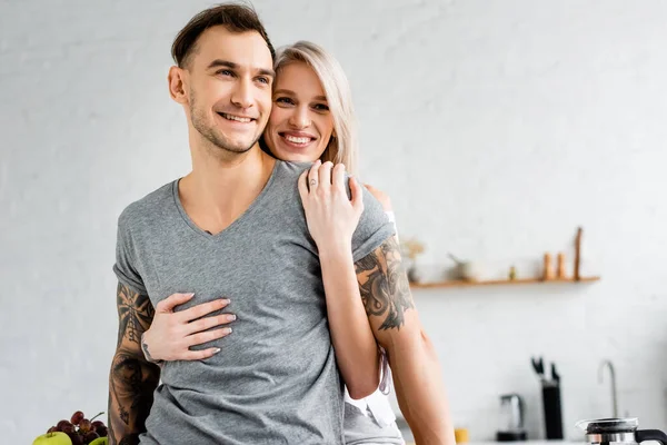 Smiling girl hugging tattooed boyfriend in kitchen — Stock Photo