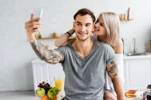 Enfoque selectivo del hombre tatuado tomando selfie con teléfono inteligente cerca de la novia sonriente en la mesa de la cocina - foto de stock