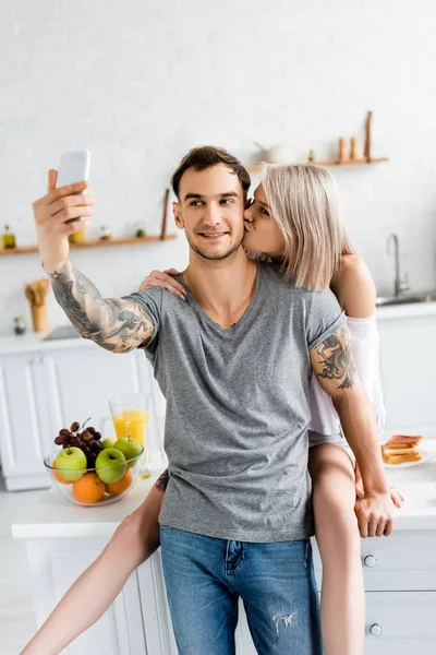 Woman kissing tattooed boyfriend taking selfie with smartphone in kitchen — Stock Photo