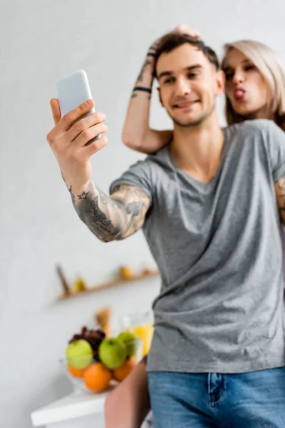 Выборочный фокус улыбающегося мужчины, делающего селфи со смартфоном рядом с татуированной девушкой на кухне — стоковое фото