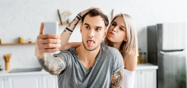 Tiro panorâmico de casal tatuado sorrindo enquanto toma selfie com smartphone na cozinha — Fotografia de Stock