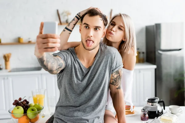 Selective focus of tattooed couple grimacing while taking selfie with smartphone during breakfast in kitchen — Stock Photo