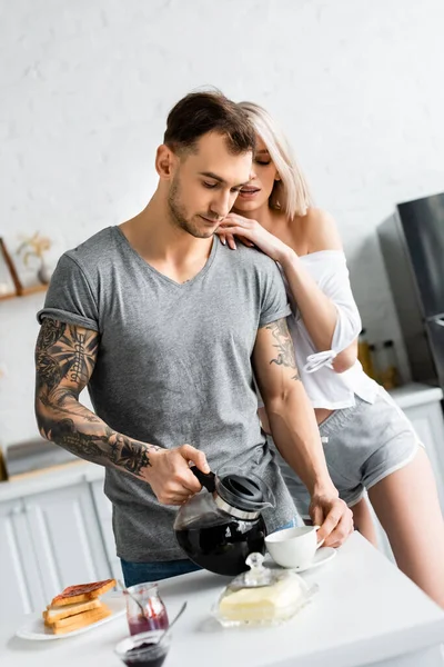 Mulher atraente abraçando bonito namorado tatuado com cafeteira perto do café da manhã na mesa da cozinha — Fotografia de Stock