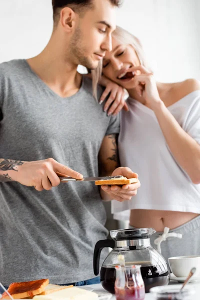 Selective focus of tattooed man with toast and knife looking at smiling girlfriend eating grape near breakfast in kitchen — Stock Photo